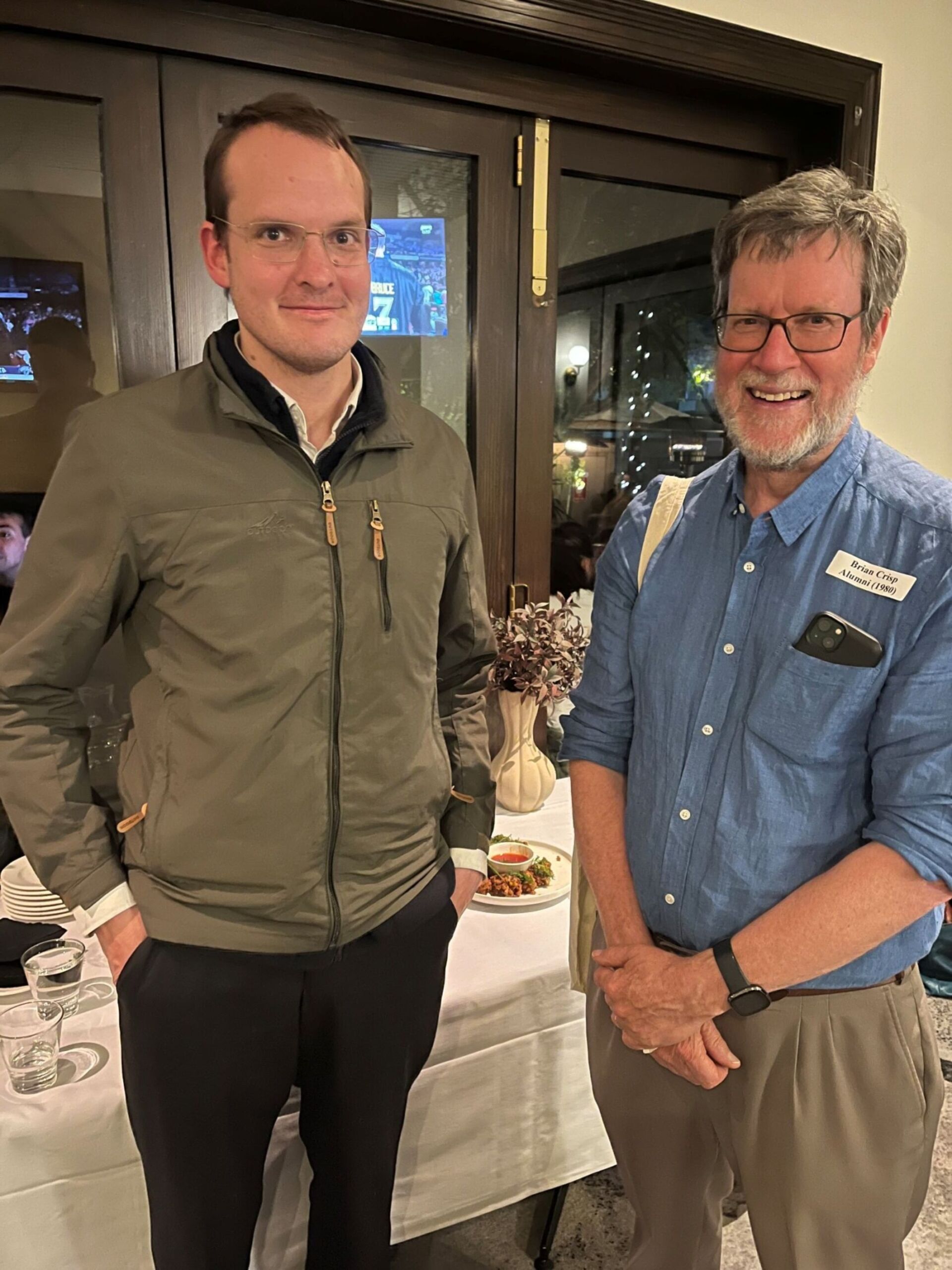 Two Burgmann alumni stand together in front of a window at the 2024 Melbourne event.