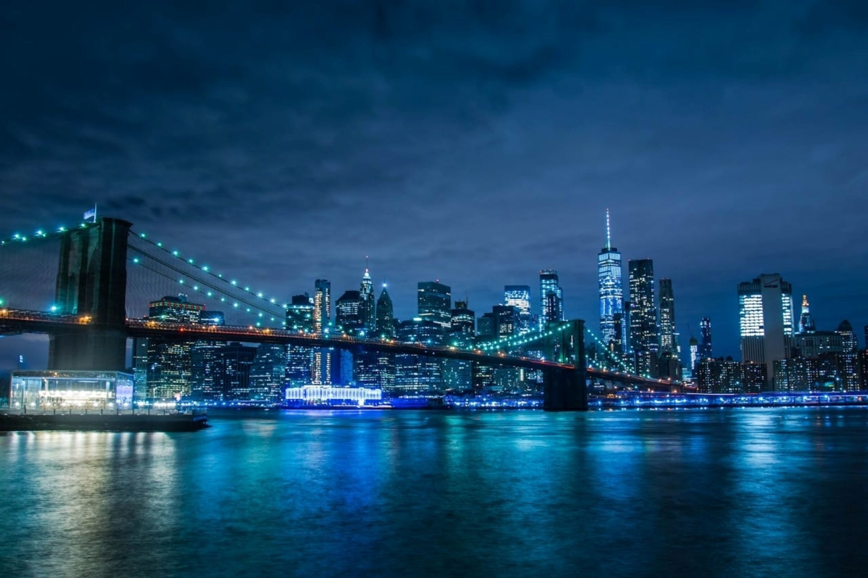 An evening photograph of the Brooklyn Bridge, New York, with street and building lights reflecting off the water.